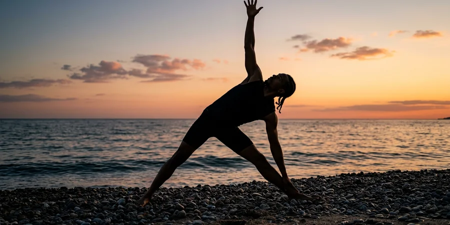 Silueta de un hombre practicando Trikonasana en una playa al atardecer, con el mar de fondo y la luz calida del sol poniente